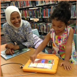 Mother in a hijab smiles as her young daughter plays on a tablet